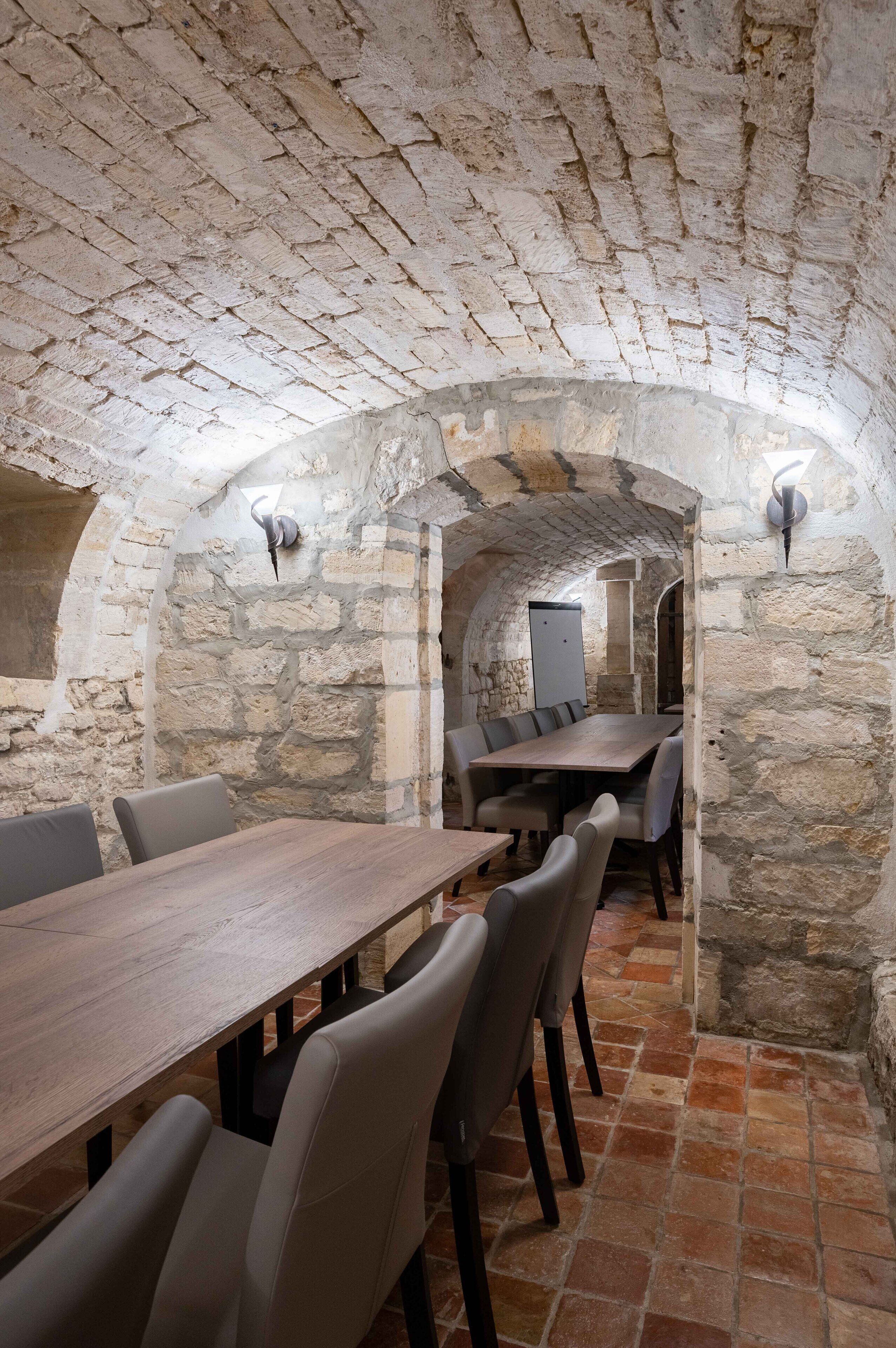 Una habitación con paredes de piedra, una mesa larga y sillas, techos abovedados y una puerta pequeña al fondo.