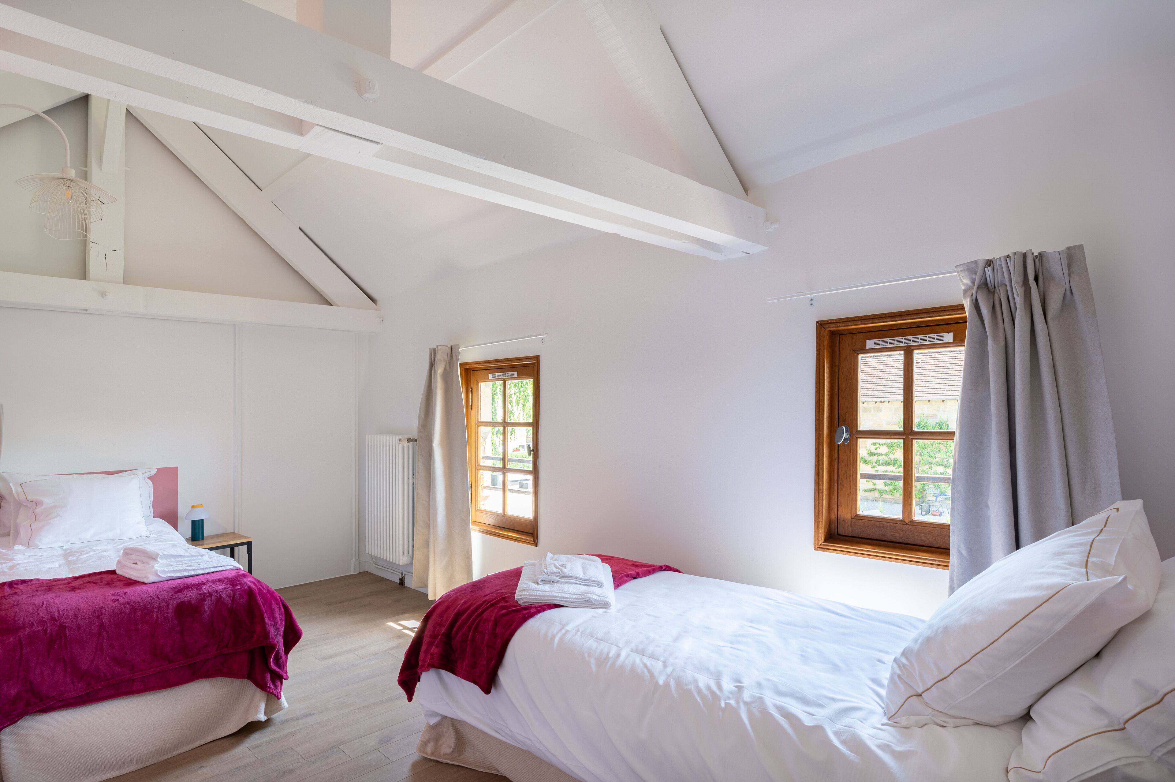 A bedroom with two beds, a window with curtains, a radiator, and a small table with a lamp.