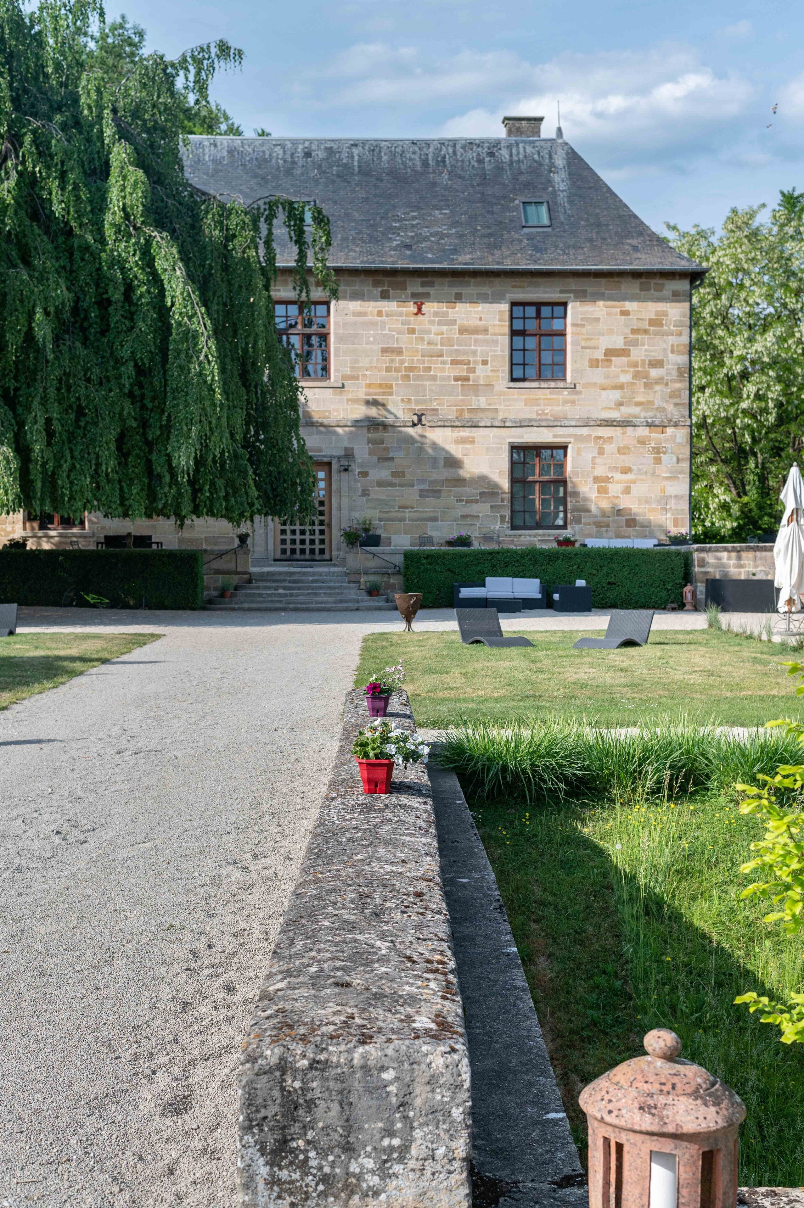 A stone building with a paved driveway, a small garden with a bench, and a pathway leading to the entrance.