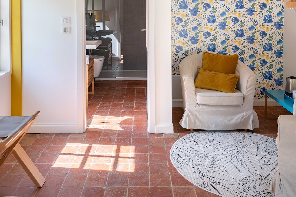 A two-story interior with a staircase, a bathroom with a shower, a dining area, and a living space with a patterned rug and a chair.