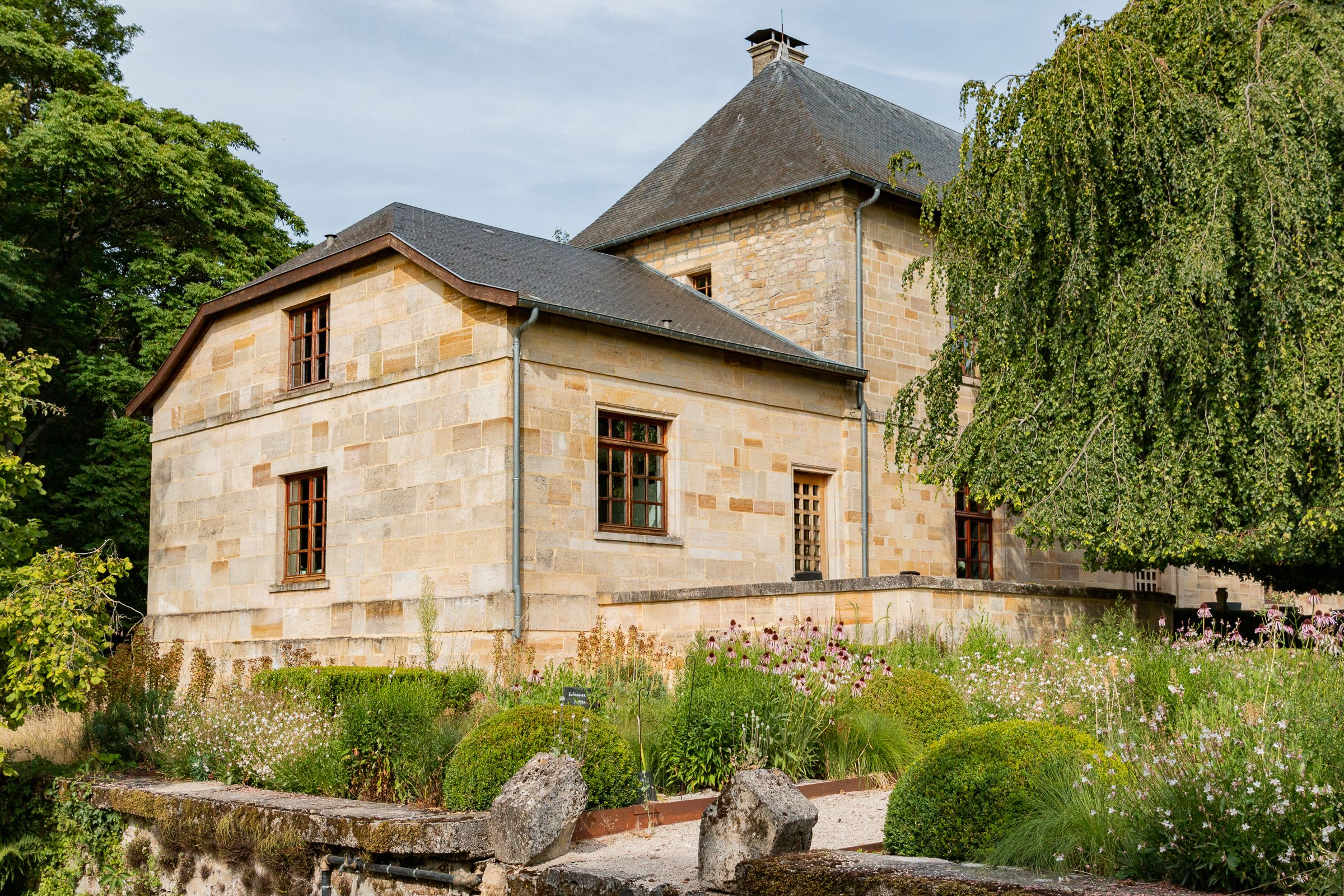 Un bâtiment en pierre doté d’un toit en pente, entouré d’une végétation luxuriante et de plantes en fleurs.