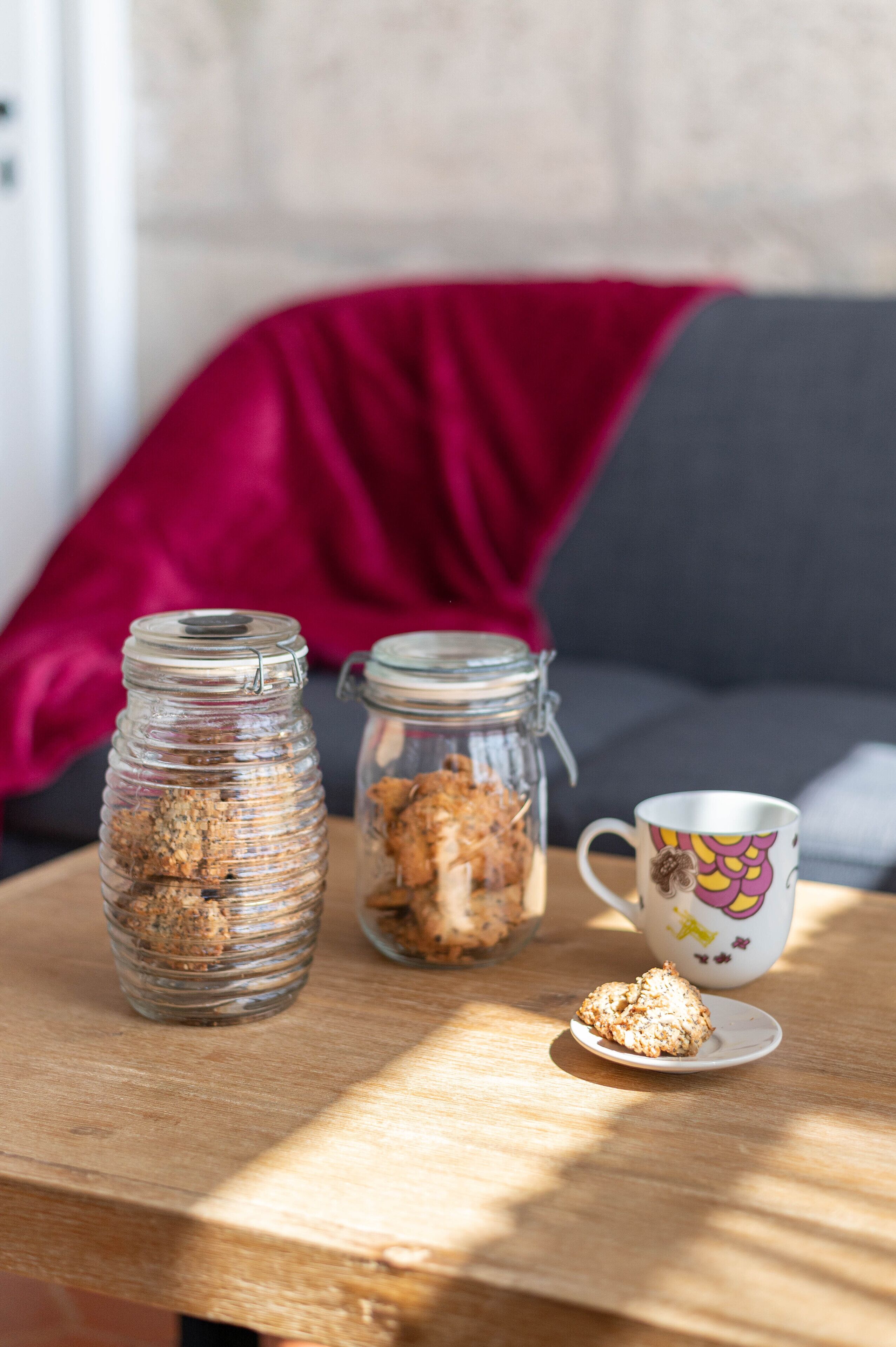 Two glass jars with contents on a wooden table, a mug with a colorful design, and a red cushion in the background.