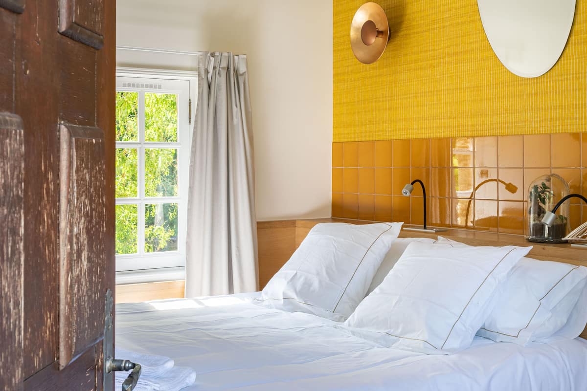 A hotel room with a bed, a window with curtains, a mirror, and a wall with a yellow pattern.