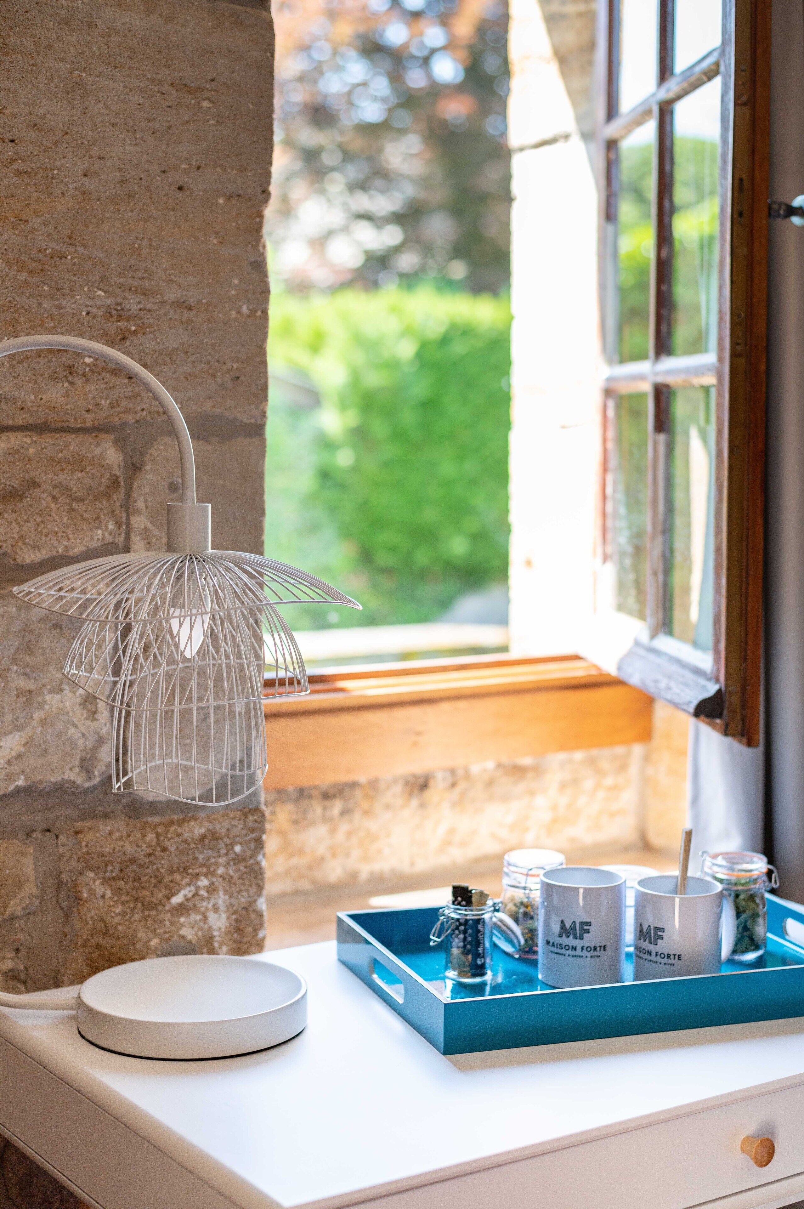 A white table with a white lamp, a blue tray with jars and mugs, and a view of greenery outside.