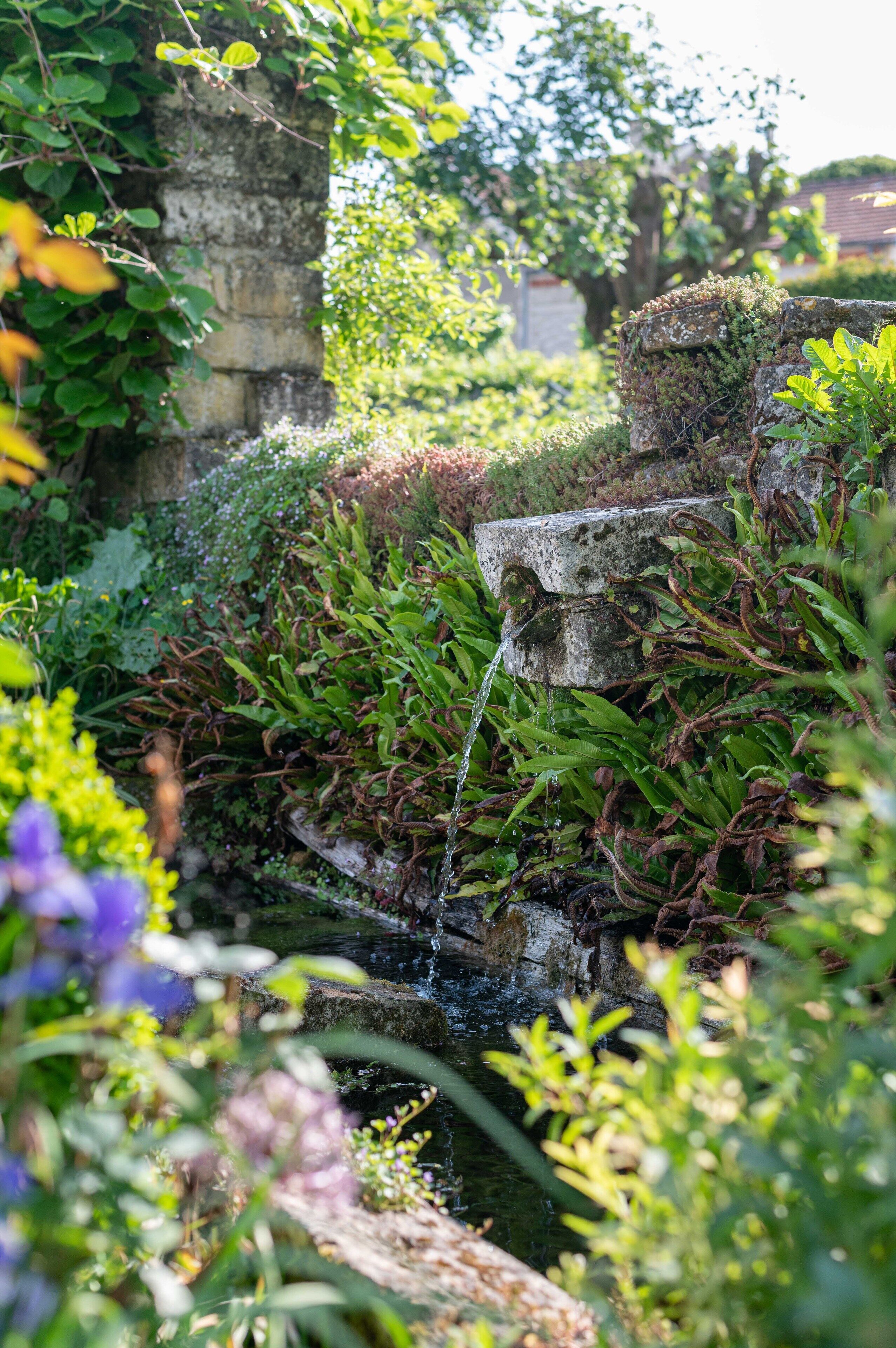 Une fontaine en pierre avec un ruisseau qui en coule, entourée d’une végétation luxuriante et de plantes en fleurs.