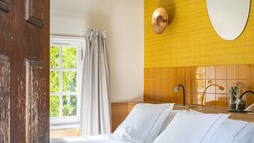 A bedroom with a bed, a wooden door, a window with curtains, and a yellow wall with a mirror.