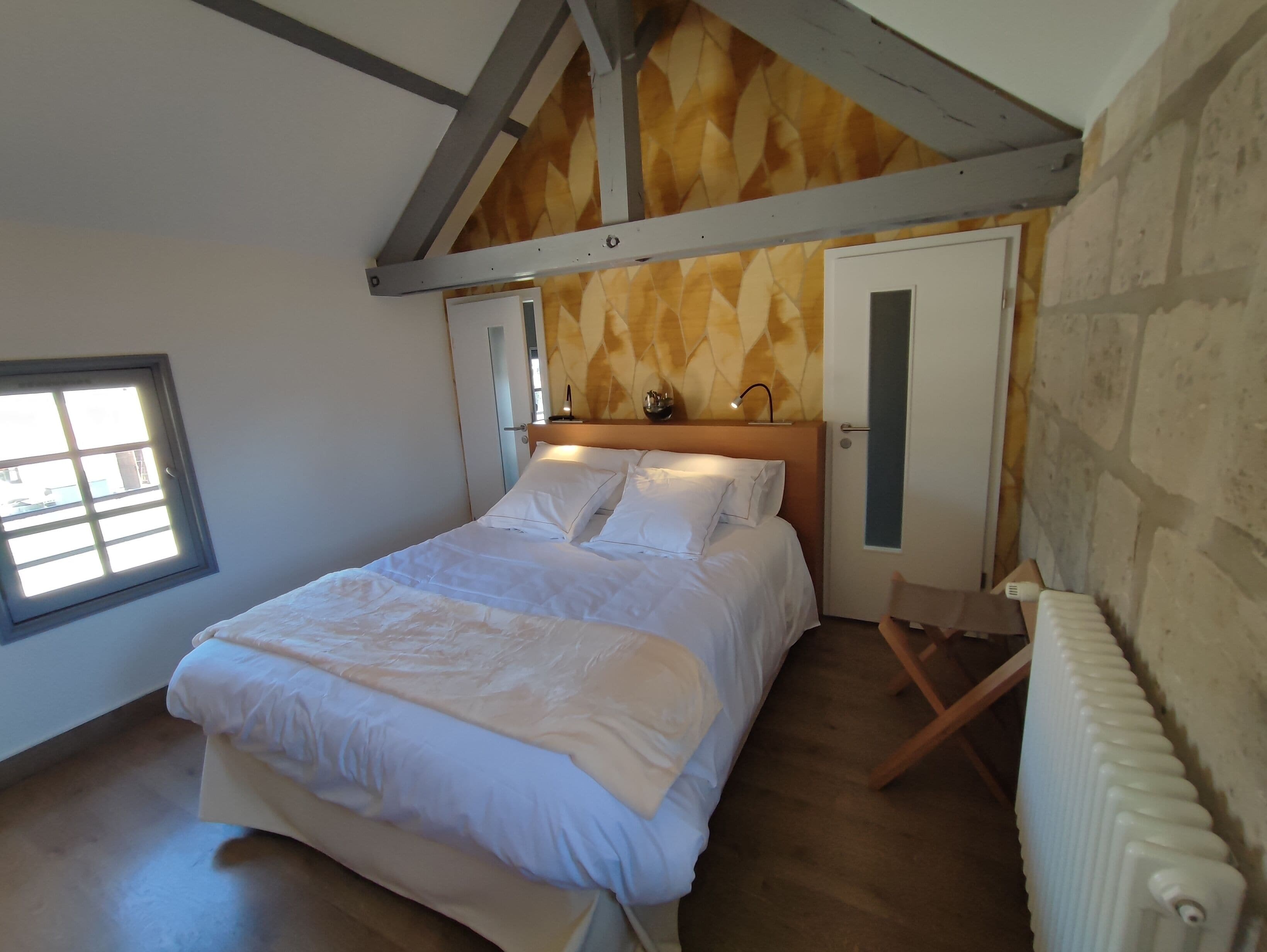 A bedroom with a wooden ceiling, a bed with white bedding, a radiator, and a window.