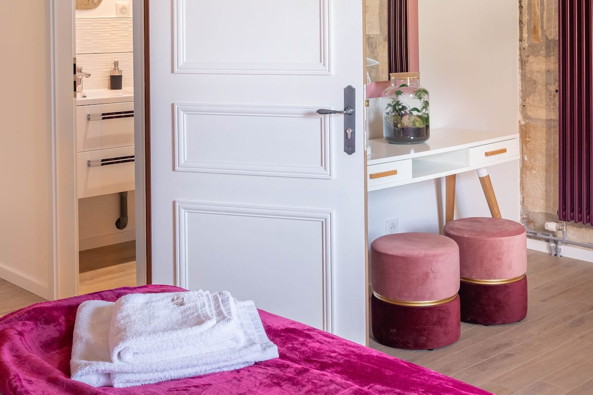 A bedroom with a bed, a pink blanket, a white towel, a white door, a vanity with a mirror, and a stone wall.