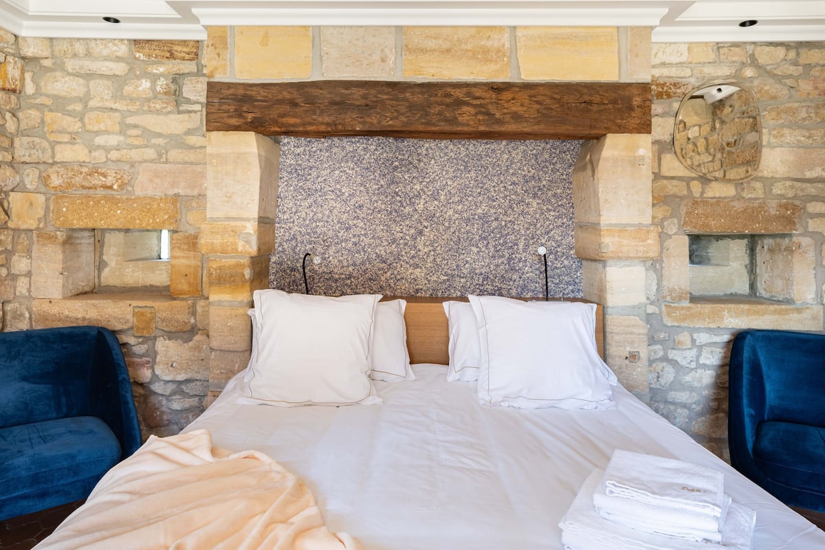 A hotel room with a stone wall, a wooden headboard, and a bed with white linens and pillows. There are two blue chairs and a stack of white towels on the bed.