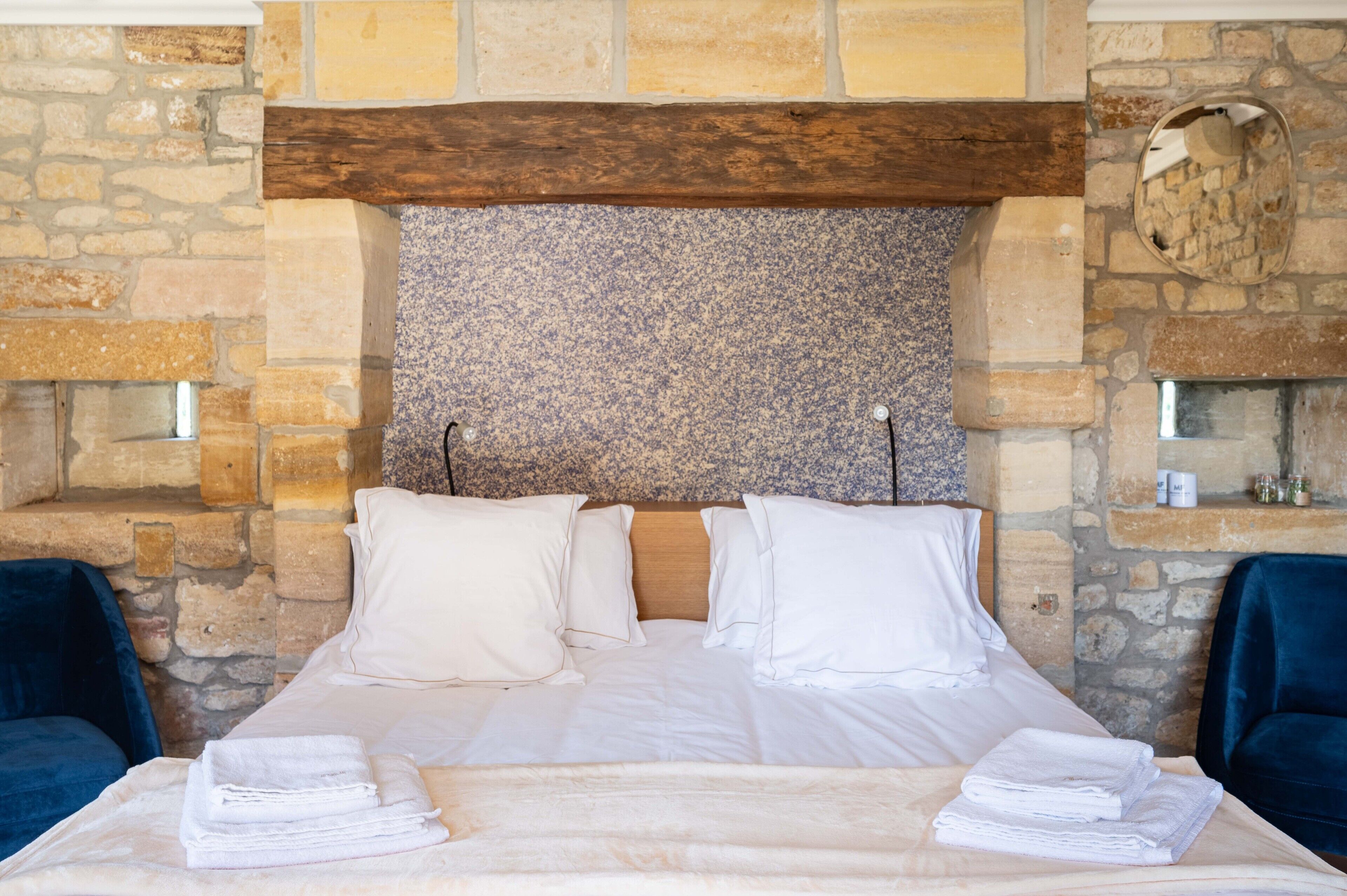 Una cama impecablemente hecha con sábanas y almohadas blancas, frente a una pared de piedra con cabecera de madera.