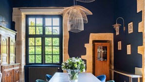 Een eetgedeelte met een lange tafel, blauwe gestoffeerde stoelen, een houten plafond en een raam met uitzicht op groen.