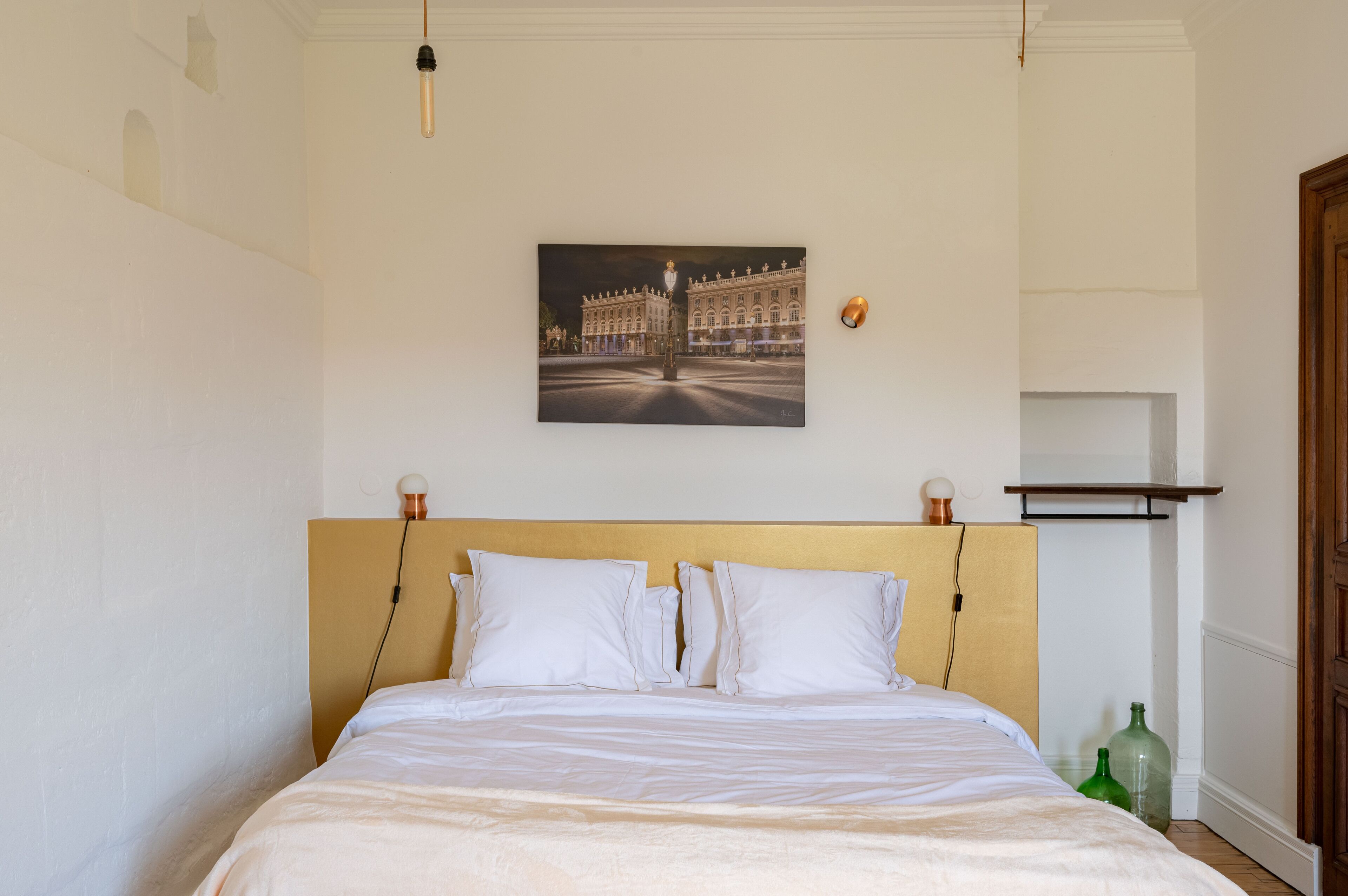 Un lit bien fait, avec du linge de lit blanc, dans une pièce dotée d’une tête de lit en bois, d’un tableau encadré au mur et d’une petite étagère avec des bouteilles.