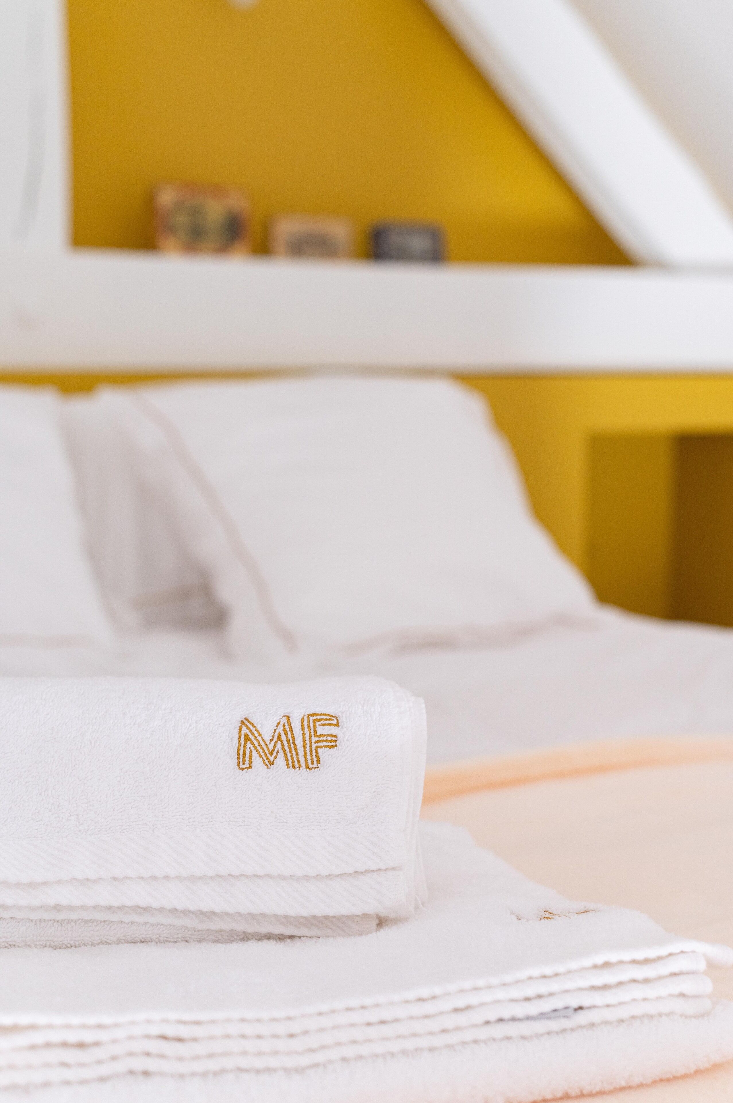 Una toalla blanca doblada con cuidado y con letras bordadas sobre una cama en una habitación con paredes amarillas.