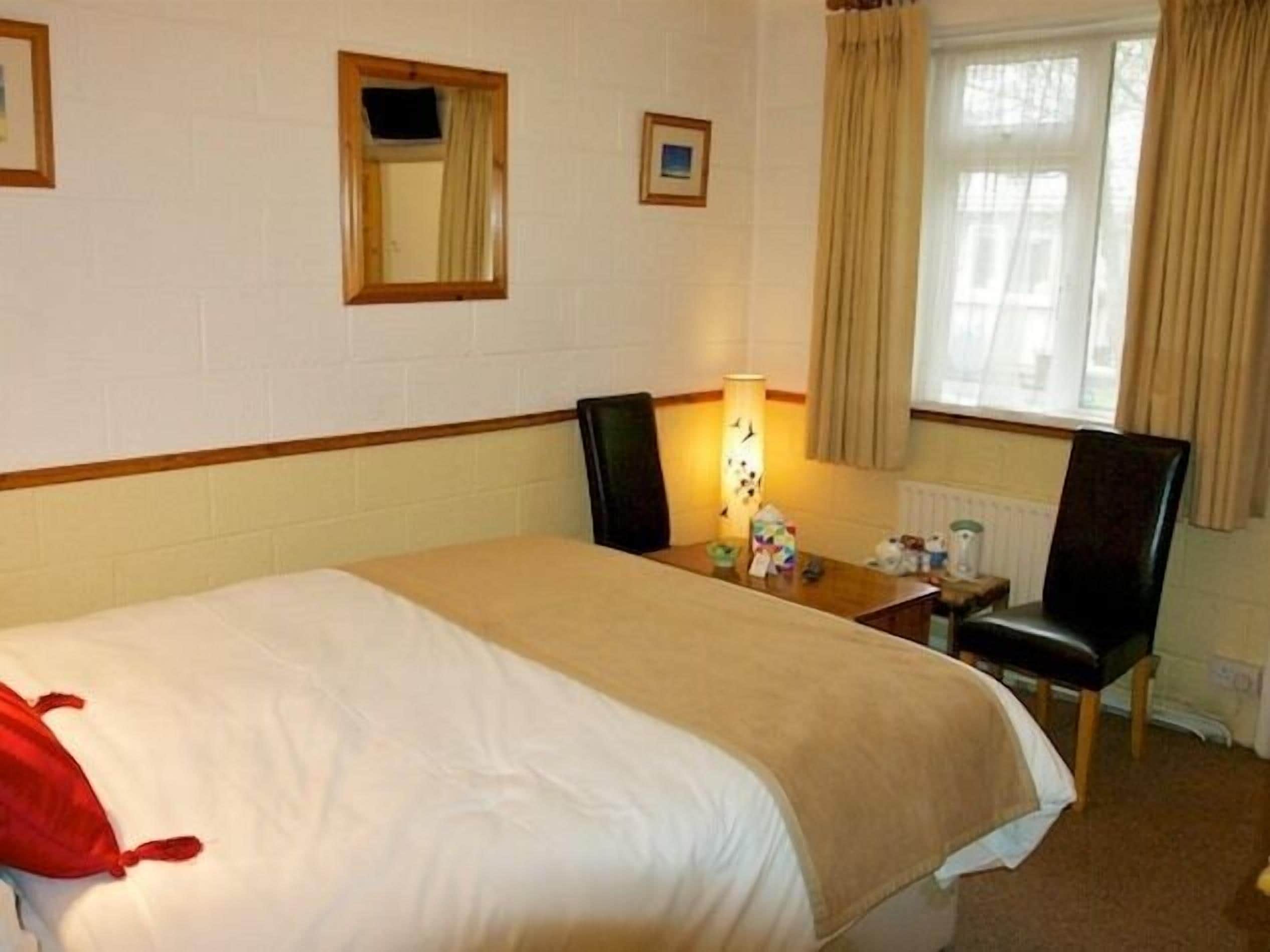 A hotel room with a large bed, a desk with a lamp, a chair, a window with curtains, and a small picture on the wall.