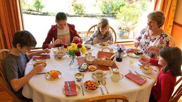 Una familia cenando en una mesa con varios platos, un jarrón de flores y vistas a un jardín por la ventana.