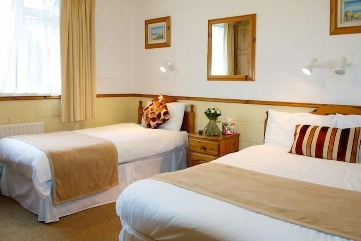 Une chambre d’hôtel avec deux lits simples, une tête de lit en bois, une table de chevet ornée d’un vase de fleurs et une fenêtre avec des rideaux.