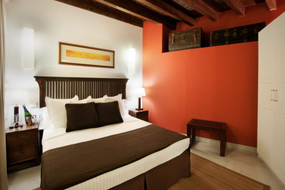 A hotel room with a large bed, wooden headboard, a nightstand with a bottle and glasses, a small table, and a red accent wall.