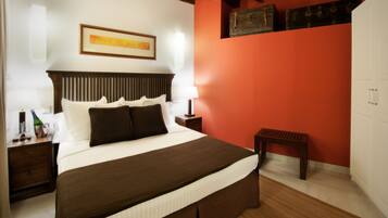 A hotel room with a large bed, wooden headboard, a nightstand with a bottle and glasses, a small table, and a red accent wall.