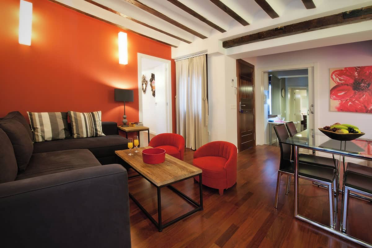 A modern living room with a dark sofa, red armchair, and wooden coffee table. A dining area with a glass table and chairs is visible in the background.