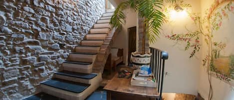 Un escalier en pierre menant à un plafond en bois, une table avec une plante et un mur orné d’œuvres d’art florales.