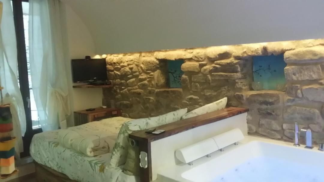 A bedroom with a stone wall, a bed with a wooden headboard, a TV mounted on the wall, and a bathtub.