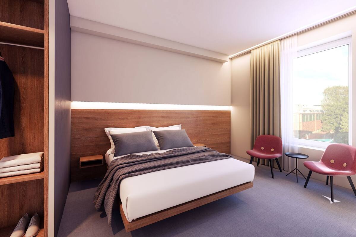 Una habitación de hotel con cama, armario, escritorio, dos sillas y una ventana con cortinas.