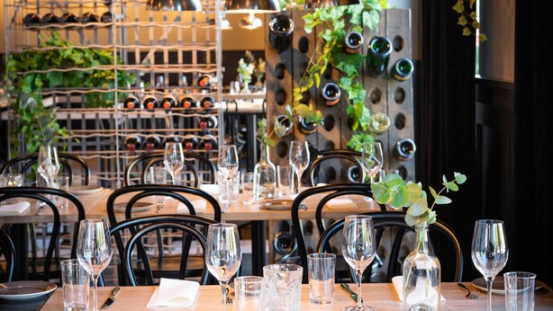 Een restaurantinterieur met gedekte tafels, wijnflessen op een rek in de achtergrond, en een muur met decoratieve flessen.