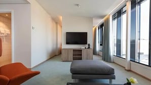 Een moderne hotelkamer met een groot raam, een flatscreen televisie, een grijze bank en een klein tafeltje met een vaas.