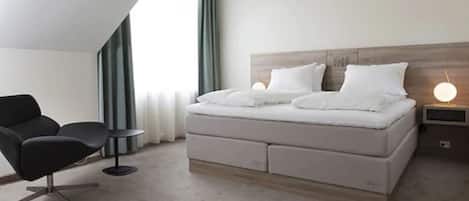 A modern hotel room with a bed, a chair, a bedside table, and a lamp.