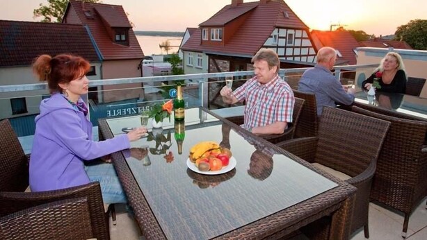 A group of people seated at an outdoor table with a glass top, surrounded by wicker chairs, with a view of houses and a sunset in the background. - Hotel Müritzperle (Waren (Müritz))