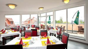 A restaurant with multiple tables, red and black chairs, and large windows offering a view of the cityscape. - Hotel Müritzperle (Waren (Müritz))