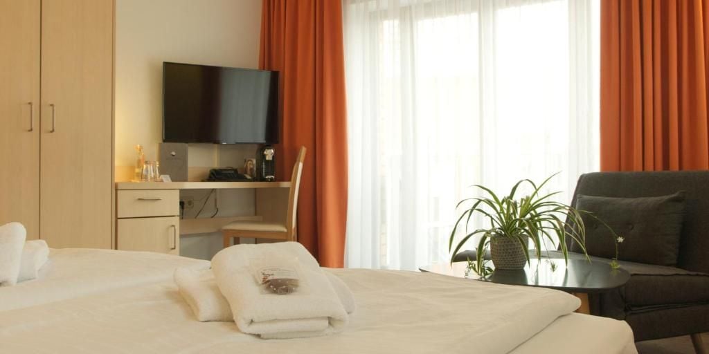 A hotel room with a bed, a desk with a flat-screen TV, a chair, a sofa, and a potted plant.