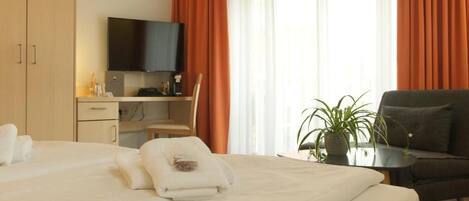 A hotel room with a bed, a desk with a flat-screen TV, a chair, a sofa, and a potted plant.