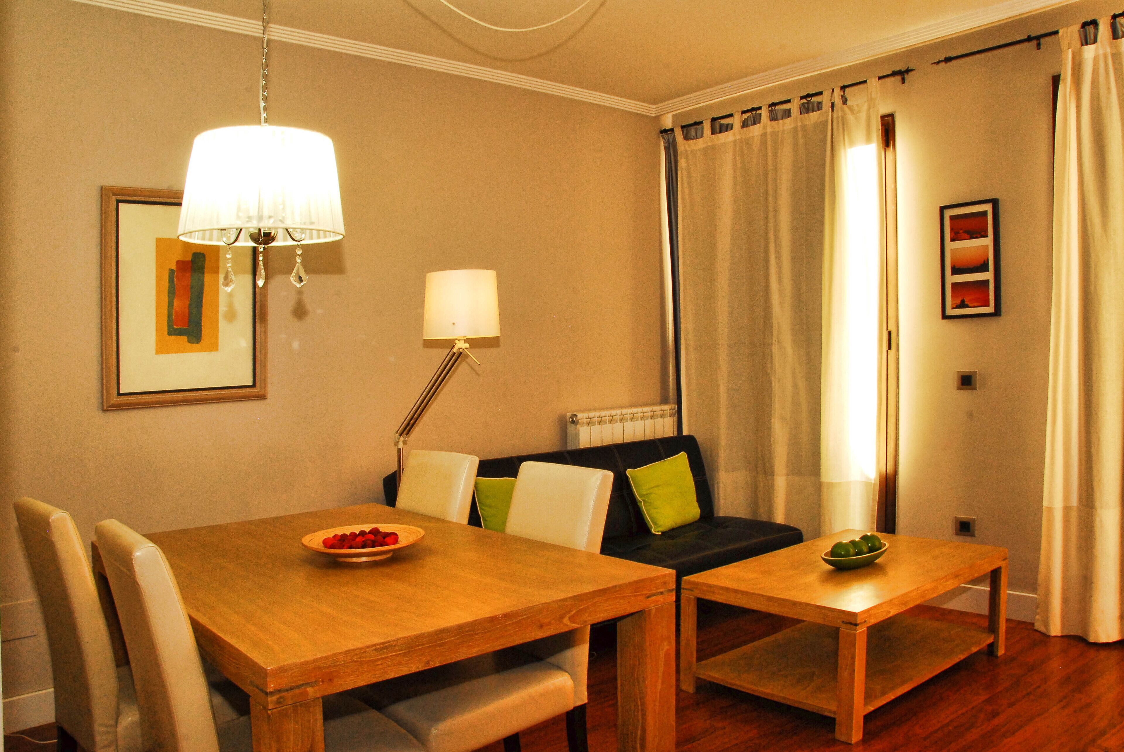 A dining area with a wooden table and chairs, a small side table with a bowl, a sofa with cushions, a floor lamp, and a framed picture on the wall.