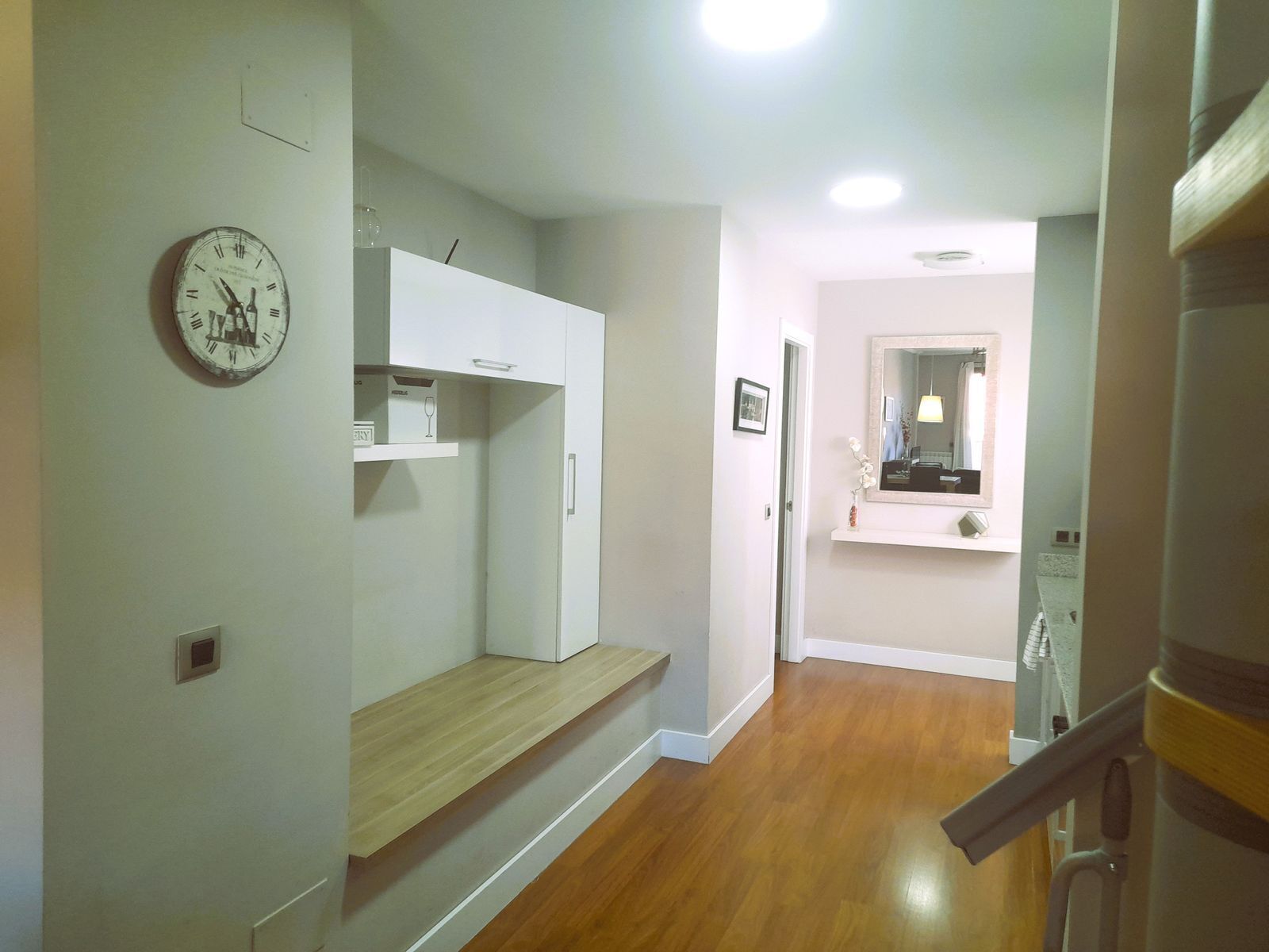 Un couloir doté d’un plancher en bois, d’une horloge murale blanche, d’une armoire blanche et d’un miroir surmonté d’un luminaire.