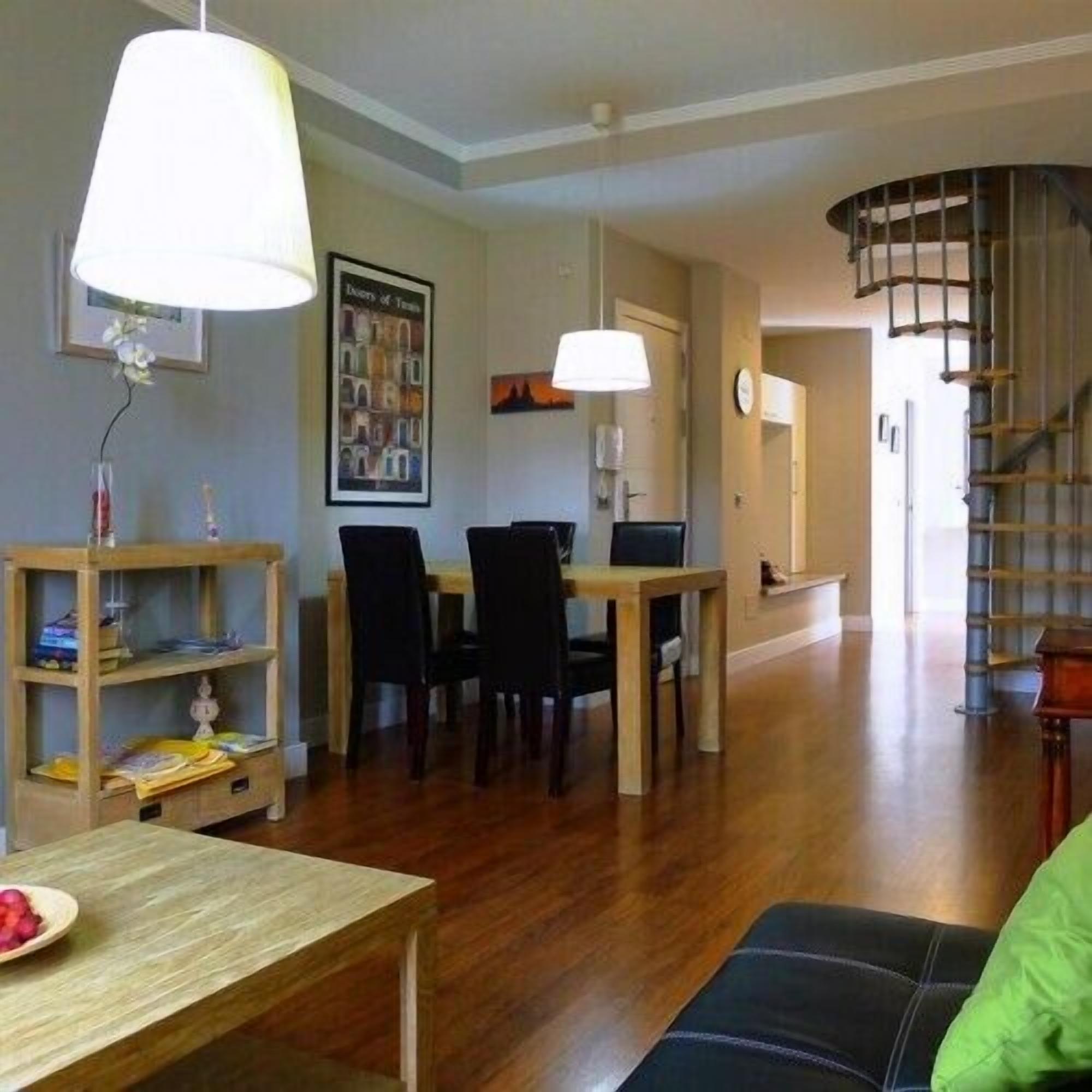 A spacious living room with a wooden dining table, chairs, a sofa, and a staircase.