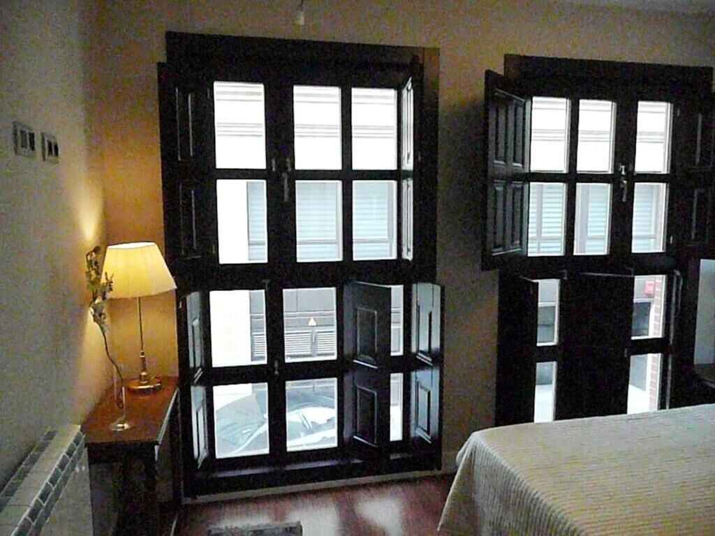 A hotel room with two large windows, a bedside table with a lamp, and a bed with a white bedspread.