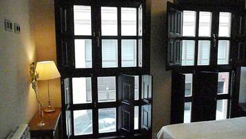A hotel room with two large windows, a bedside table with a lamp, and a bed with a white bedspread.
