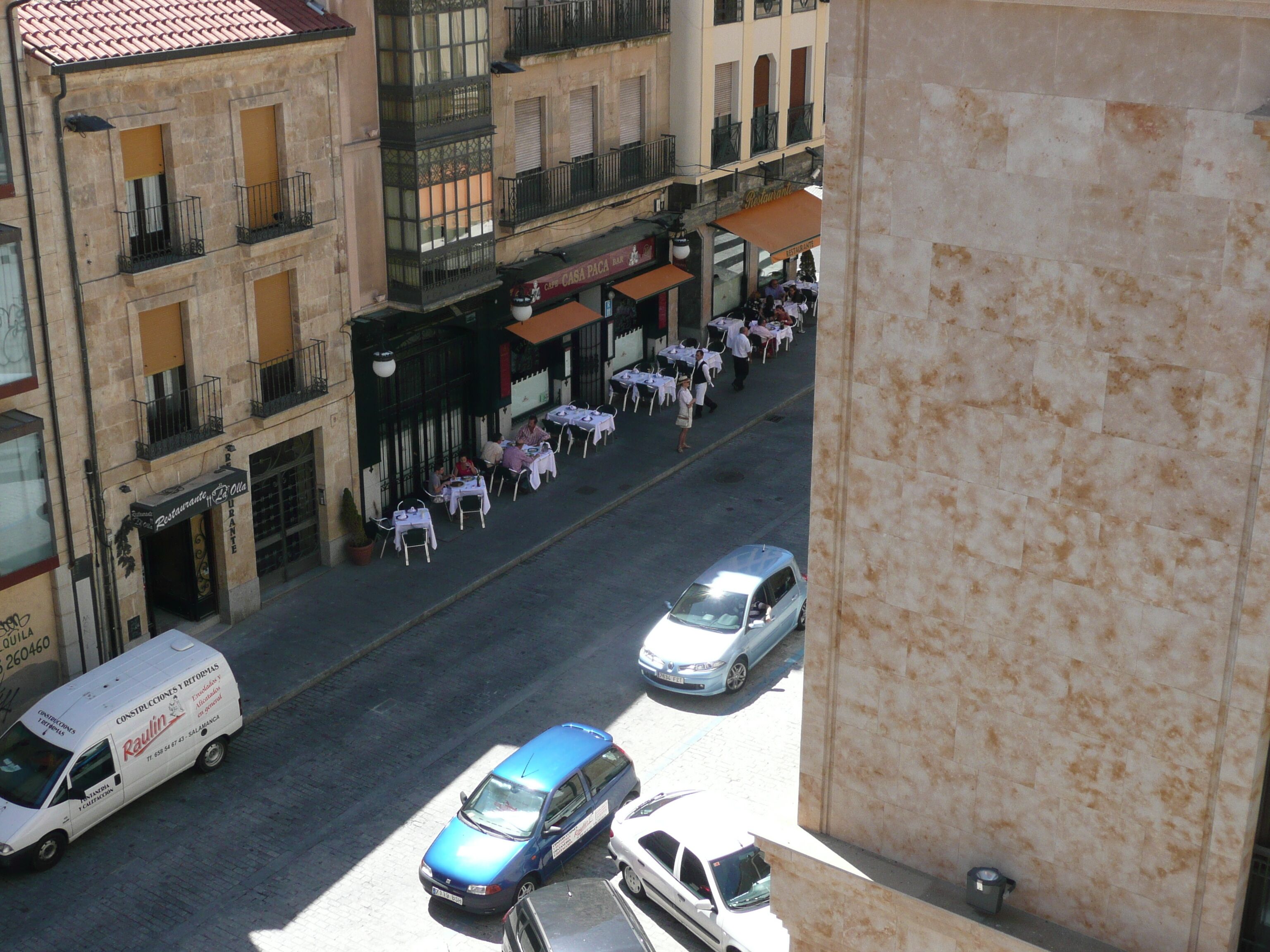Vista de la calle con terraza en un restaurante, una furgoneta de reparto estacionada y un coche azul.