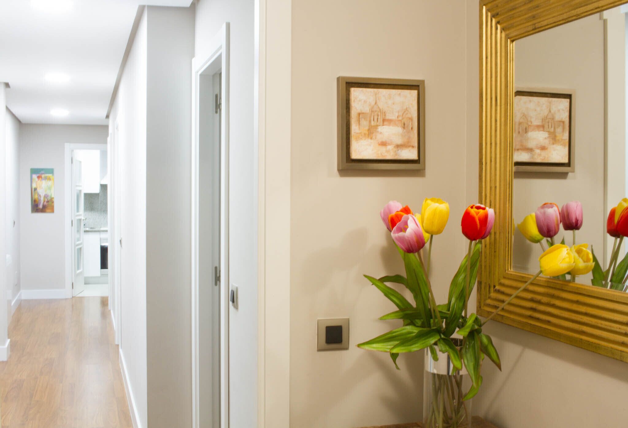 A hallway with a mirror reflecting colorful tulips, a framed picture on the wall, and a door leading to another room.