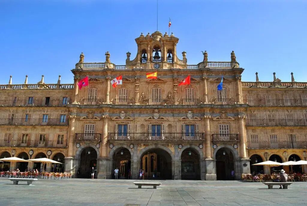 Un bâtiment historique doté de nombreuses arcades et orné de drapeaux, avec une tour horloge centrale et une cour intérieure aménagée avec des sièges en plein air.