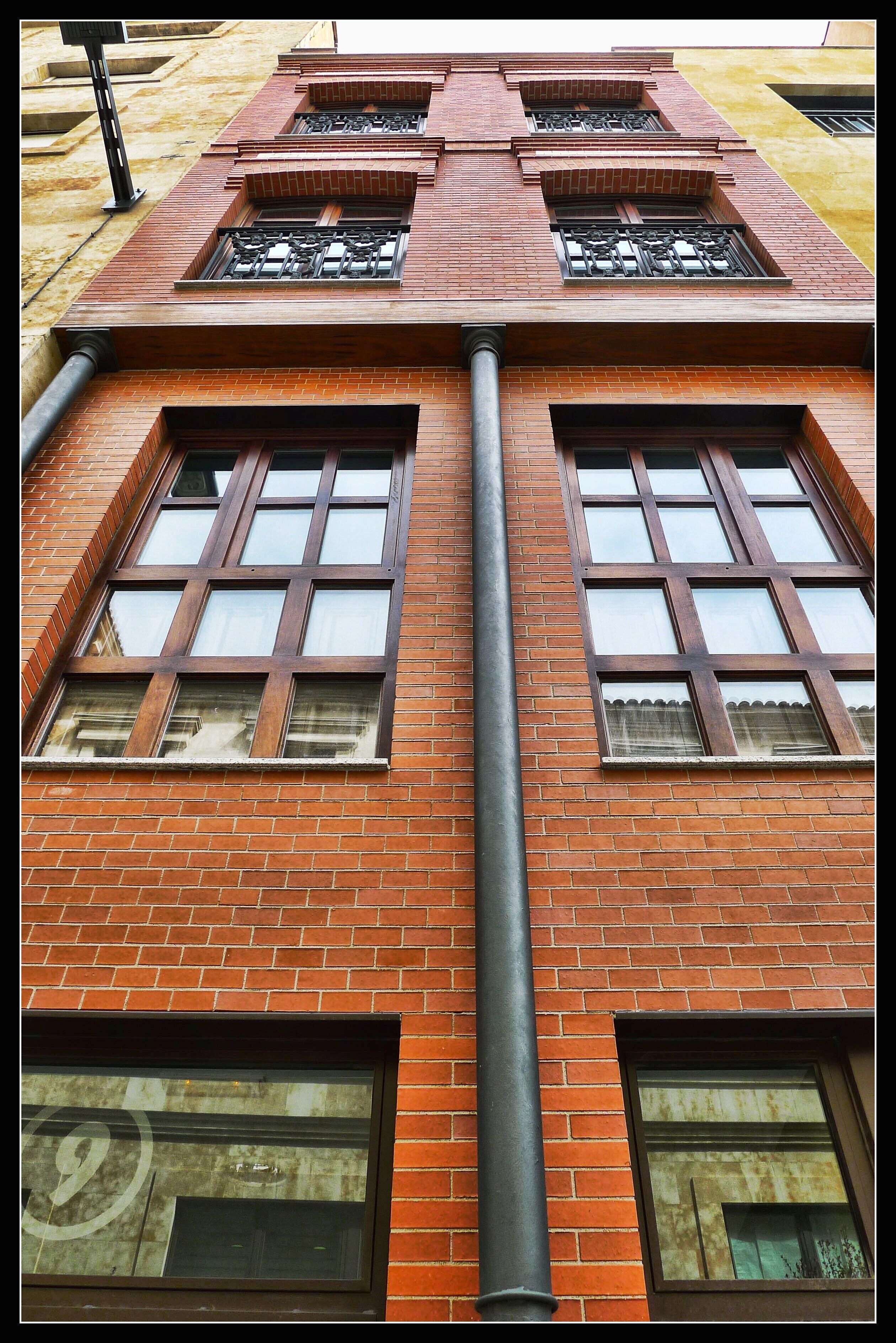 Ein rot backsteiner Gebäude mit mehreren Fenstern und einer vertikal verlaufenden schwarzen Rohrleitung.