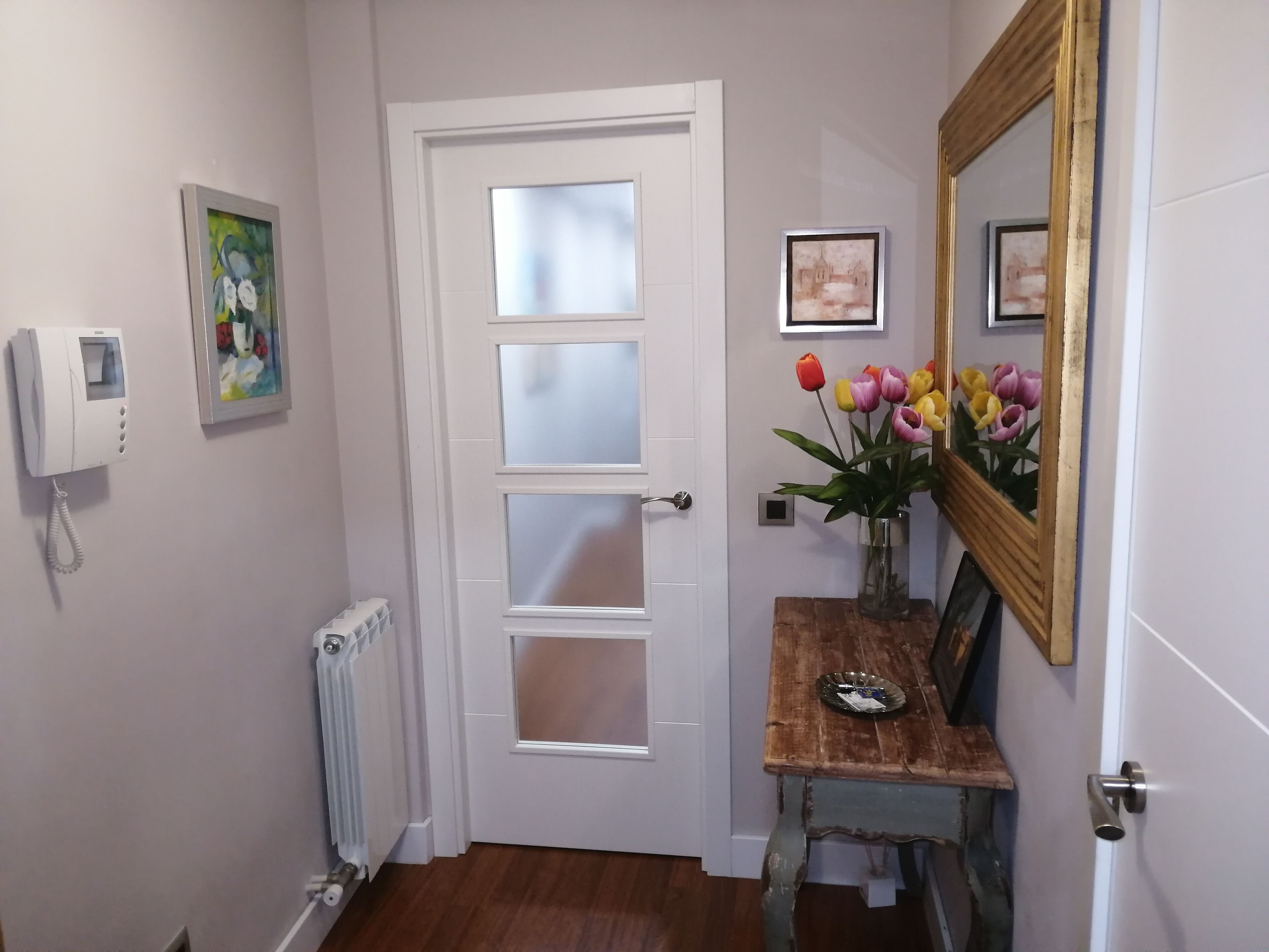 A hallway with a white door, a telephone, a radiator, a wooden table with a vase of tulips, and framed pictures on the wall.