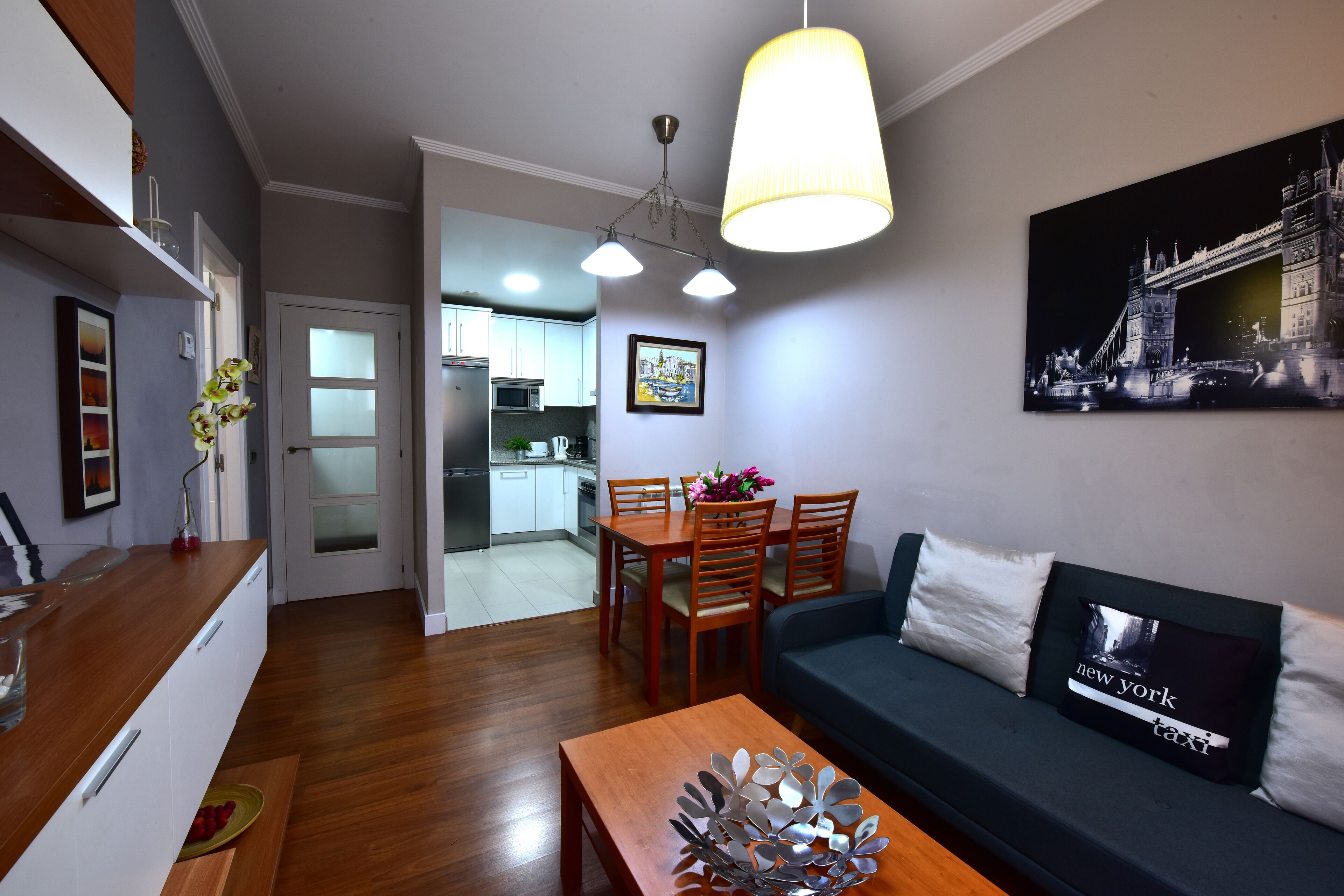 A modern living room with a wooden dining table, a sofa with decorative pillows, and a kitchen in the background.