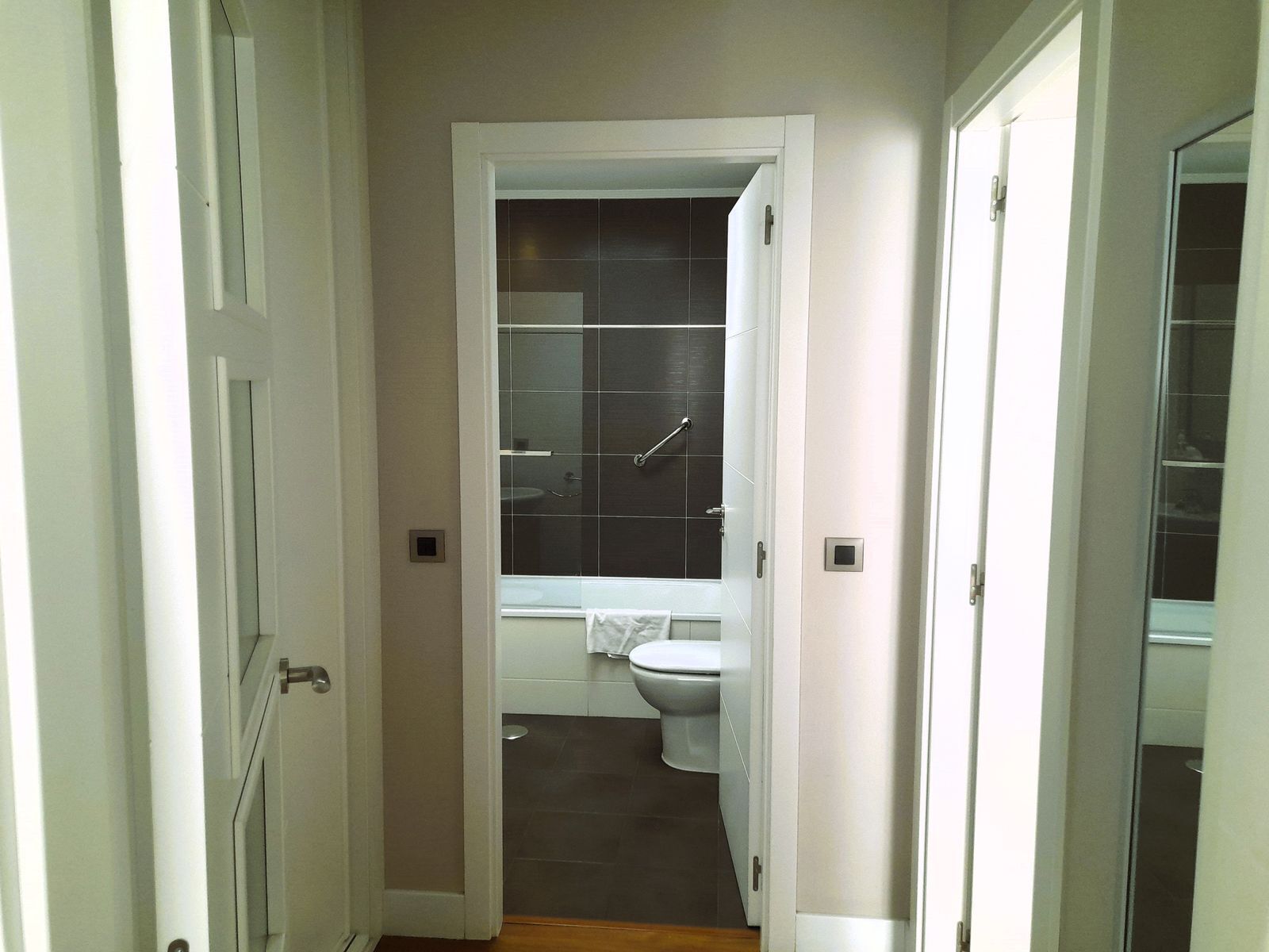 A narrow hallway leading to a bathroom with a toilet, sink, and glass shower enclosure.