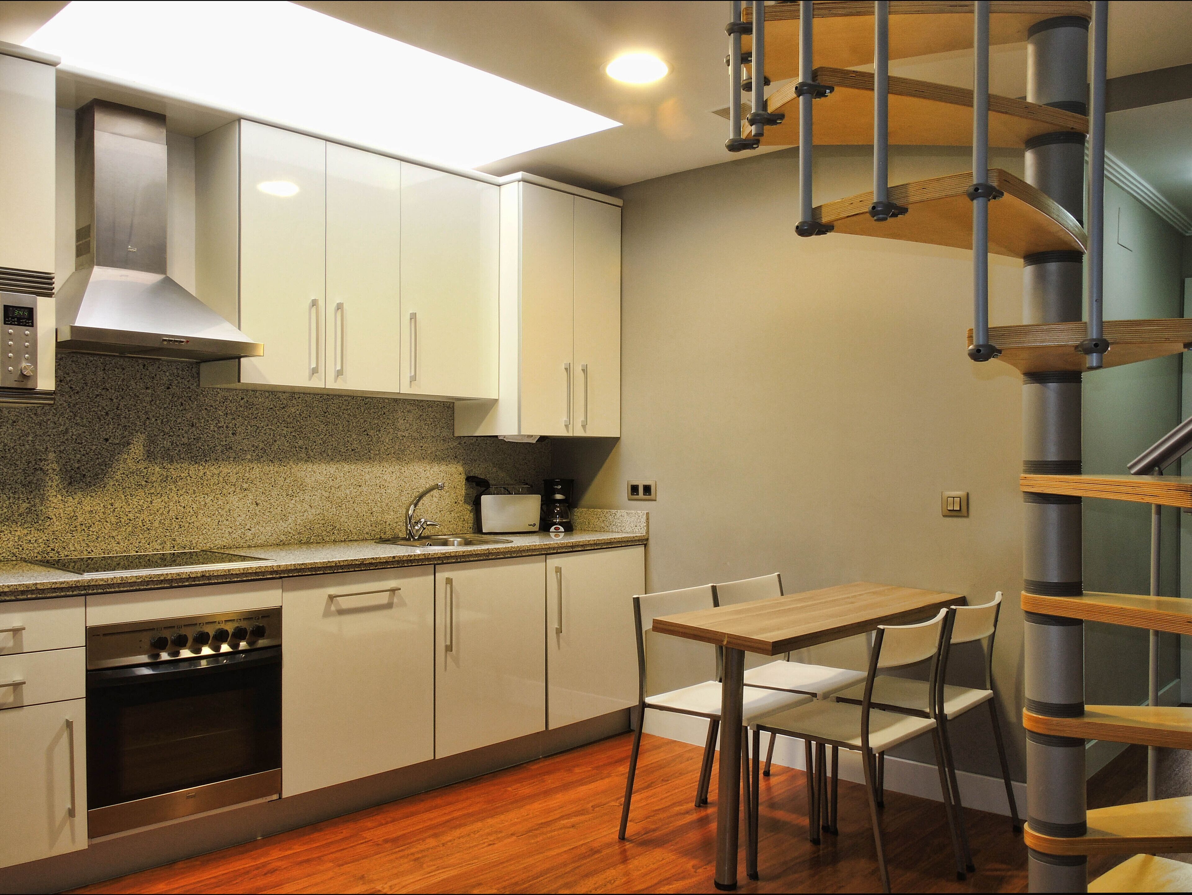 Una cocina moderna con gabinetes blancos, horno integrado, extractor de acero inoxidable y una mesa de comedor de madera con sillas.