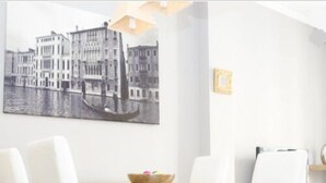 Een eetruimte met een houten tafel en witte stoelen, een zwart-wit stadsgezichtschilderij aan de muur, en een klein houten bijzettafeltje met een schaal fruit.