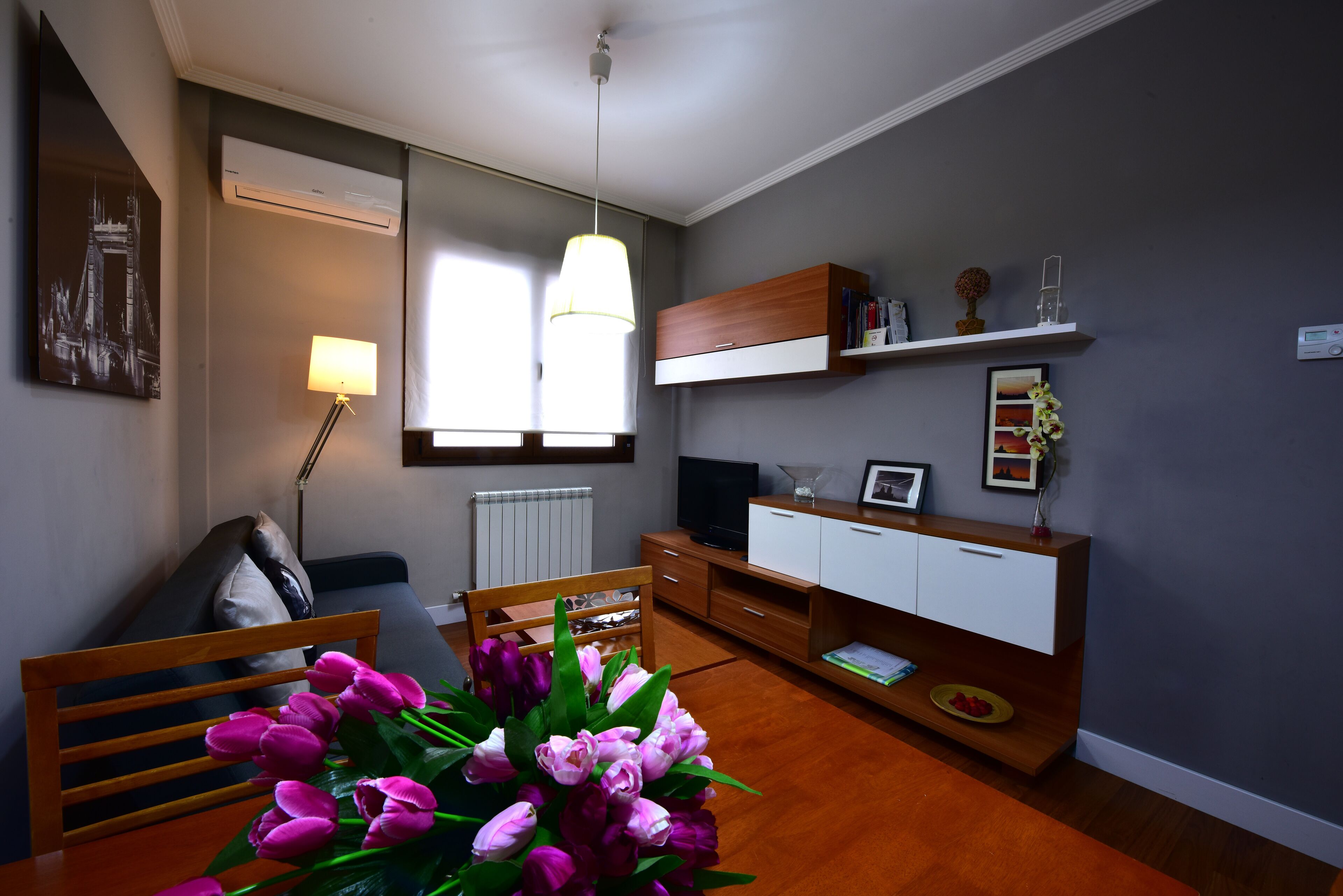 Un salon moderne avec un canapé en bois, une table à manger agrémentée d’un bouquet de tulipes, un téléviseur, une bibliothèque et une fenêtre laissant entrer la lumière naturelle.