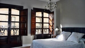 Een slaapkamer met een groot bed, een kroonluchter en uitzicht door geopende luiken.