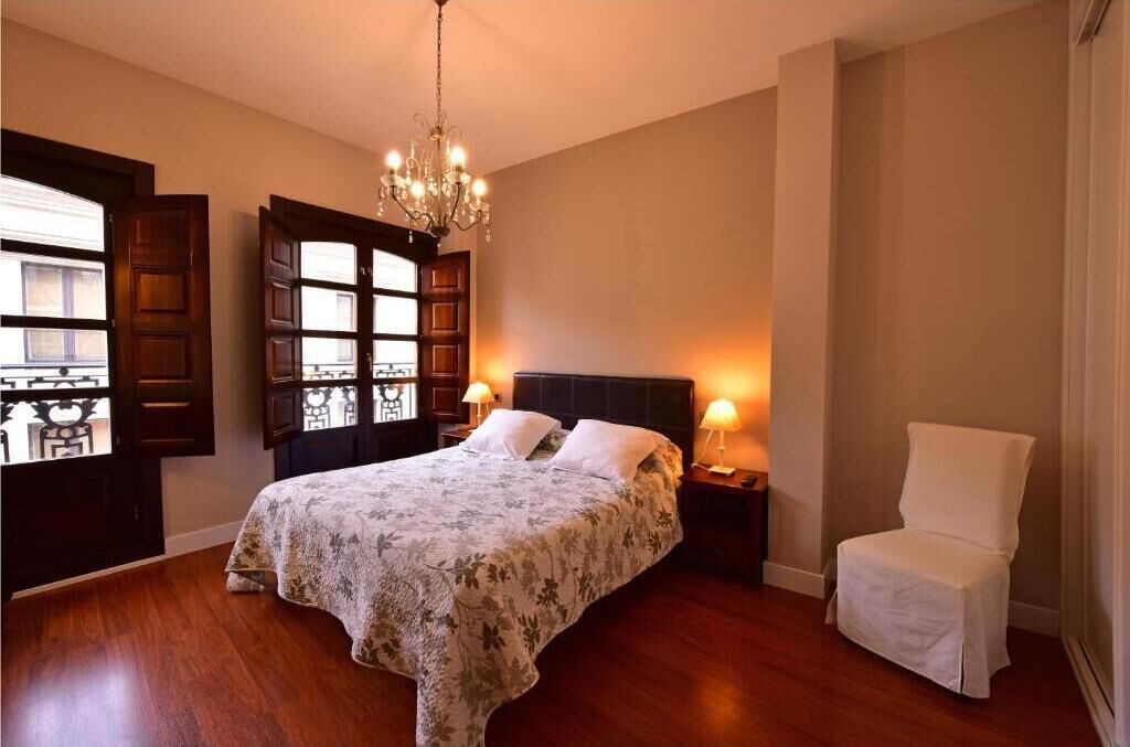 Une chambre à coucher avec un grand lit, un lustre, une chaise et une fenêtre avec des volets en bois.